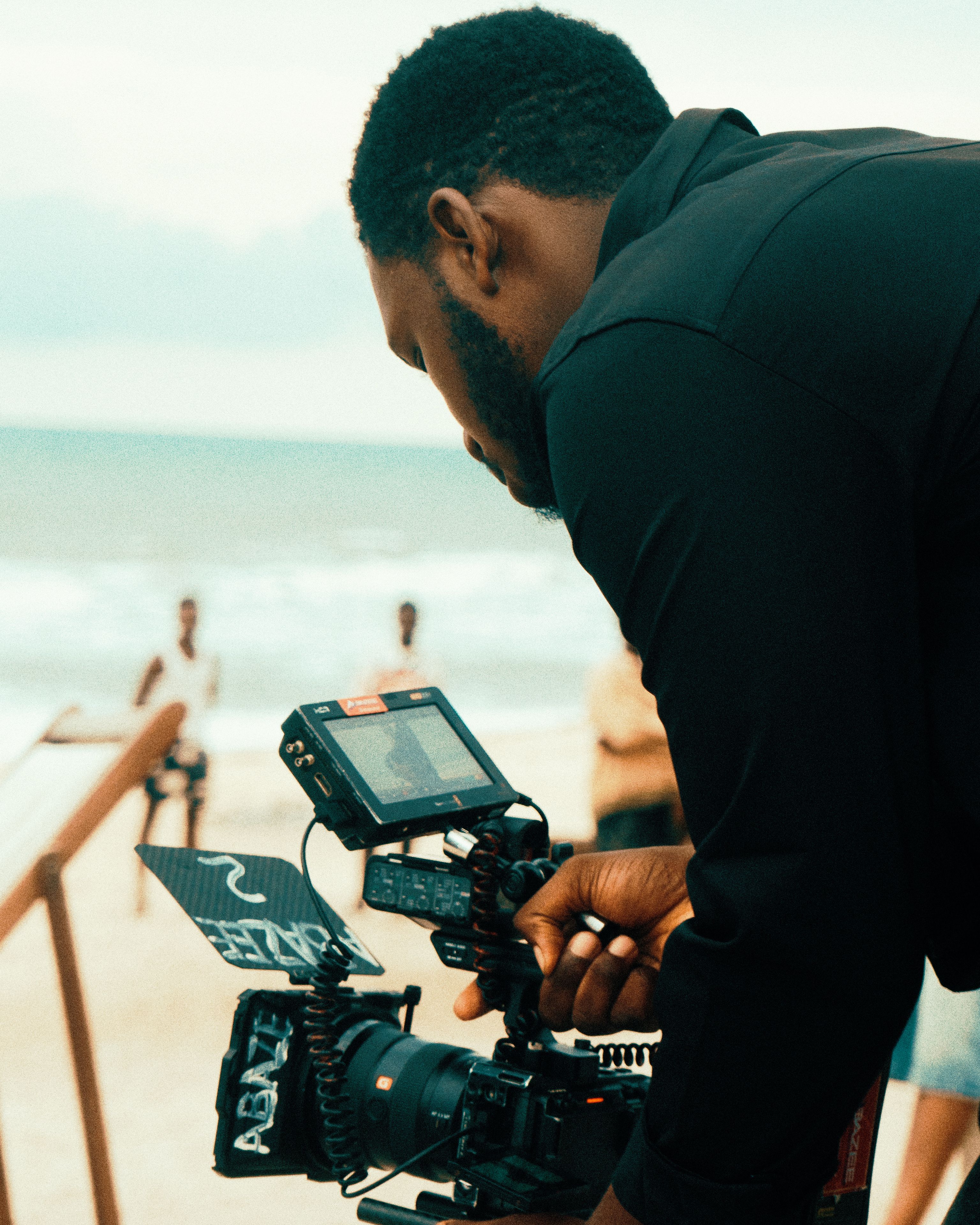 Film director adjusting camera setup on a beach during daytime shoot.