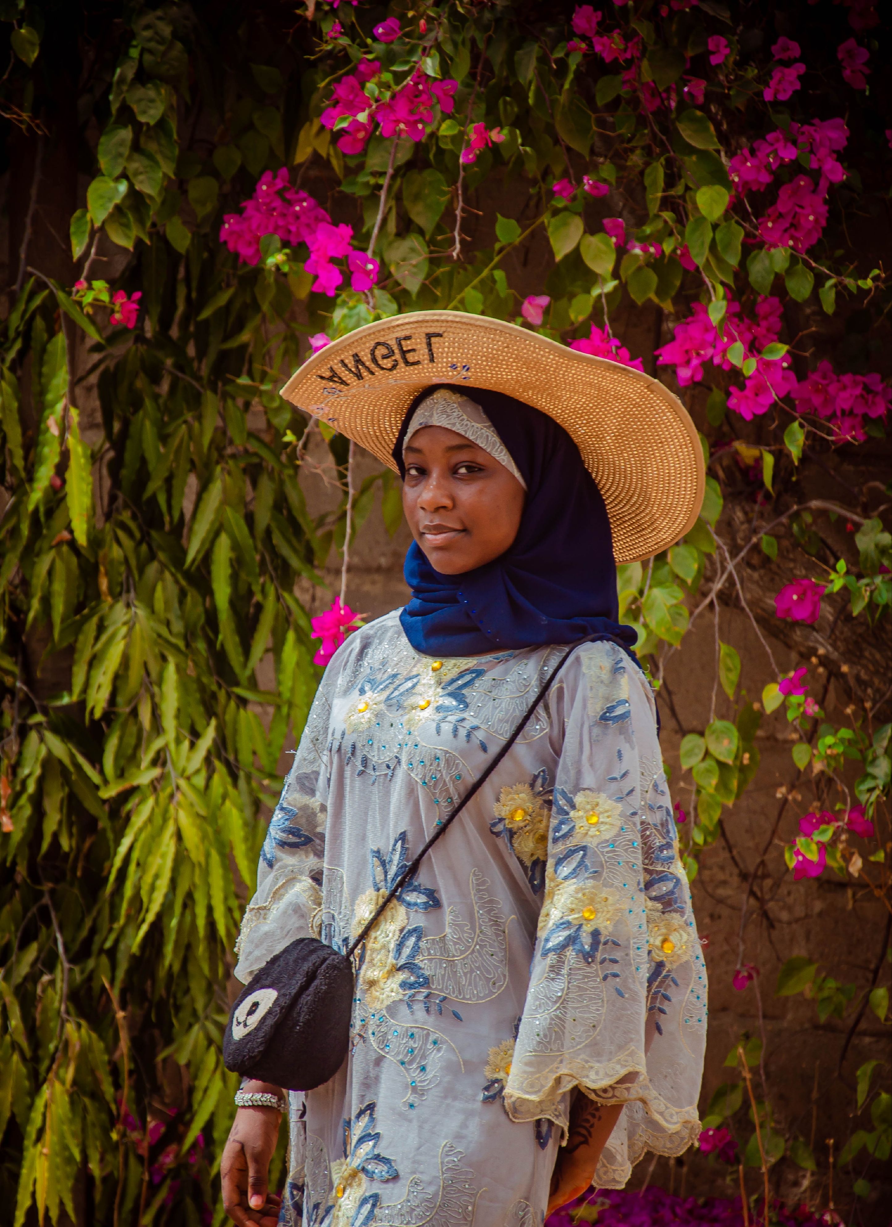 A woman wearing a hijab and straw hat stands against vibrant floral background.