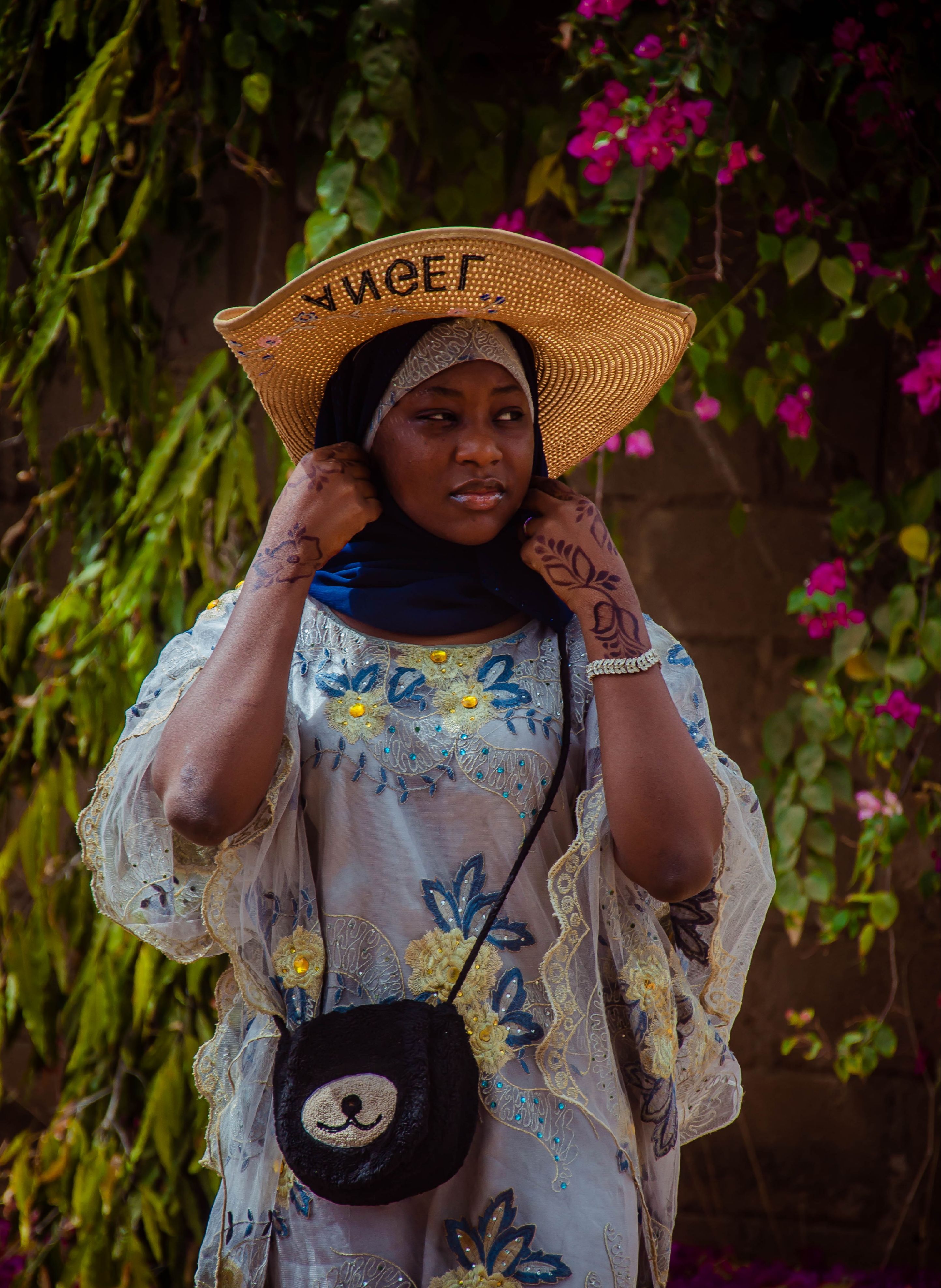 A woman wearing a straw hat and traditional dress stands amidst a floral backdrop.