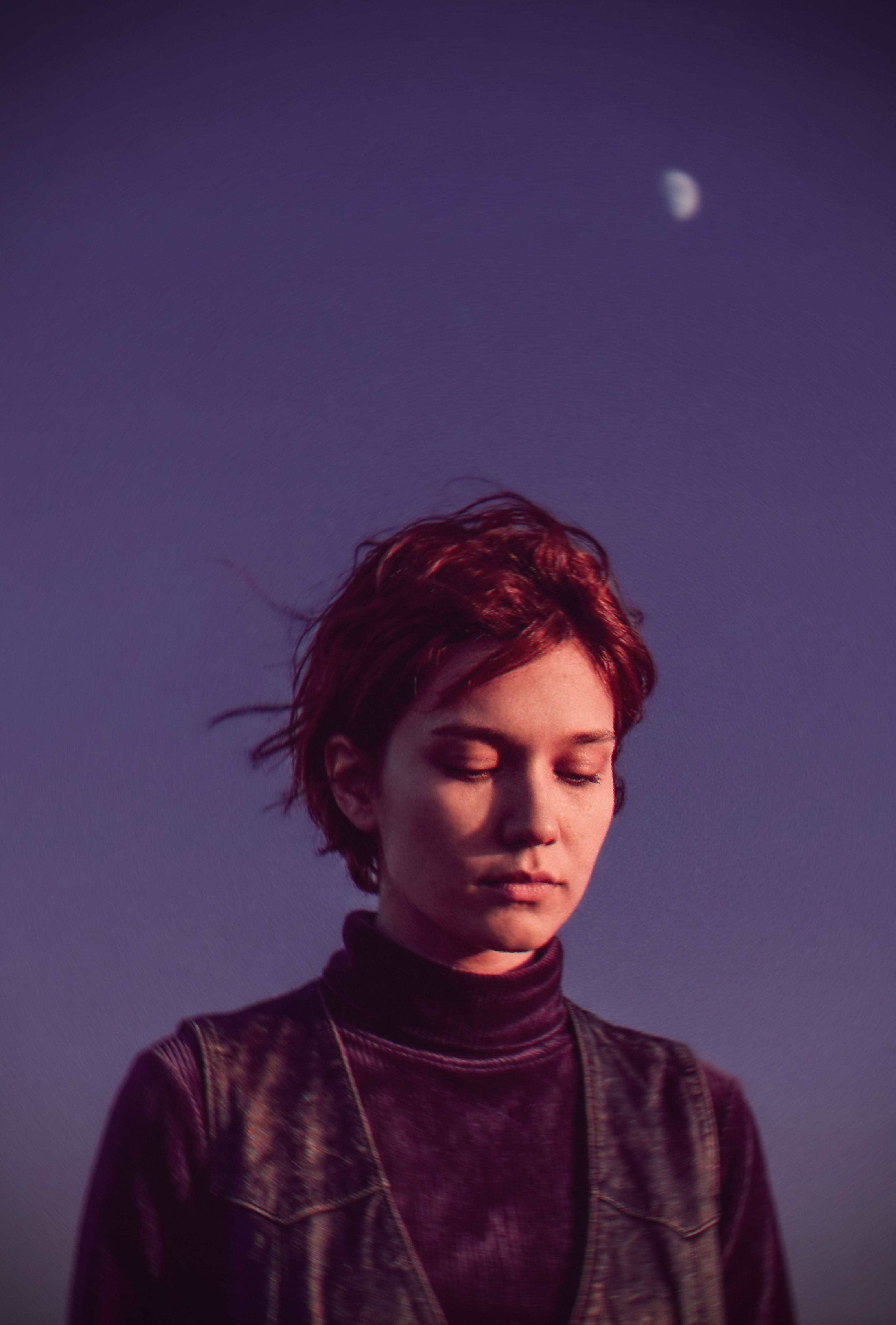 A redheaded woman with short hair and closed eyes stands under a moonlit purple sky, evoking calmness.