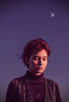 A redheaded woman with short hair and closed eyes stands under a moonlit purple sky, evoking calmness.