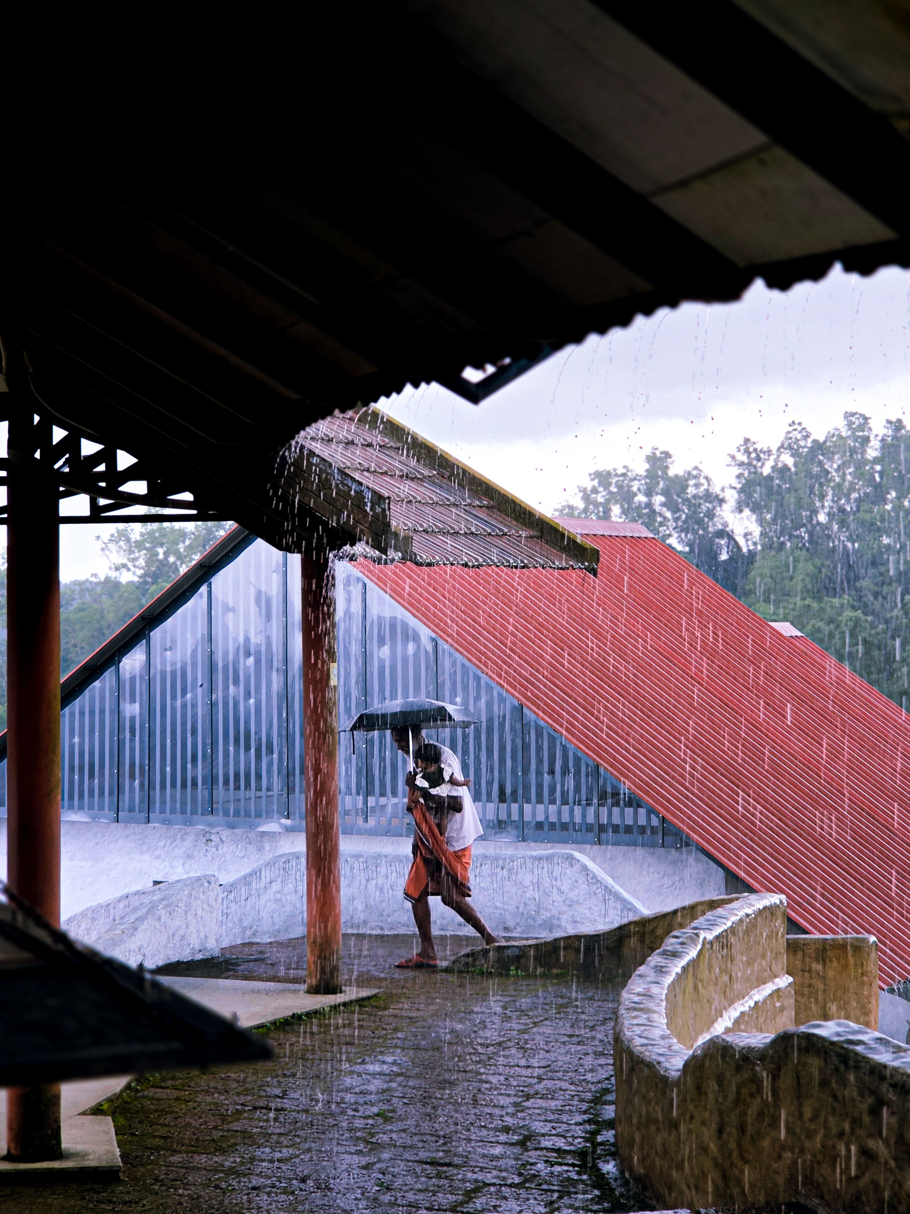 A man walks with an umbrella amidst heavy rain under traditional architectural structure in Kerala.