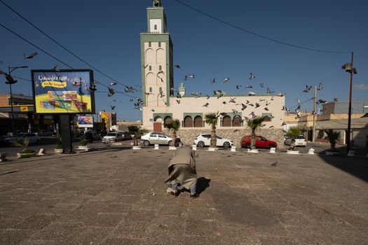 A man feeds pigeons in a lively square with a mosque in the background, Orán, Algeria.