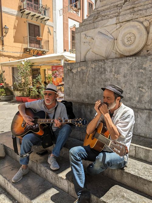 Free Two street musicians play guitars in a sunlit Italian piazza, creating a lively atmosphere. Stock Photo