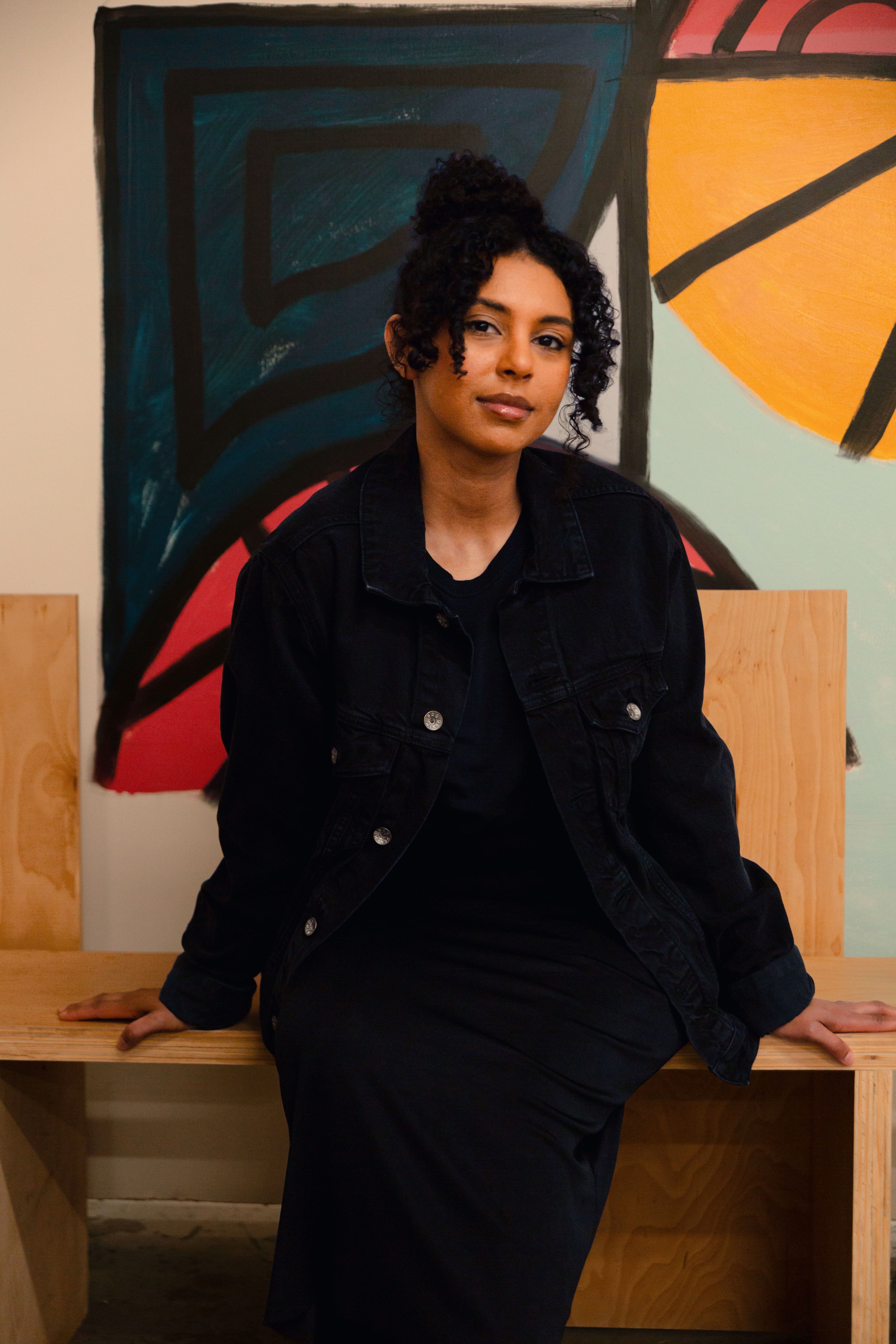 Stylish woman sitting indoors against a vibrant abstract painting, showcasing modern art and fashion.
