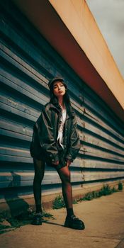 Stylish young woman in urban outfit poses against a corrugated metal wall, showcasing modern street fashion.