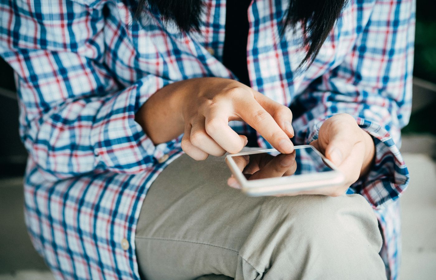 Woman's hands using a smartphone, wearing plaid shirt, indoors. Close-up shot highlighting touch interaction.