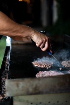 Close-up of hands grilling beef patties on a flat griddle outdoors during nighttime.