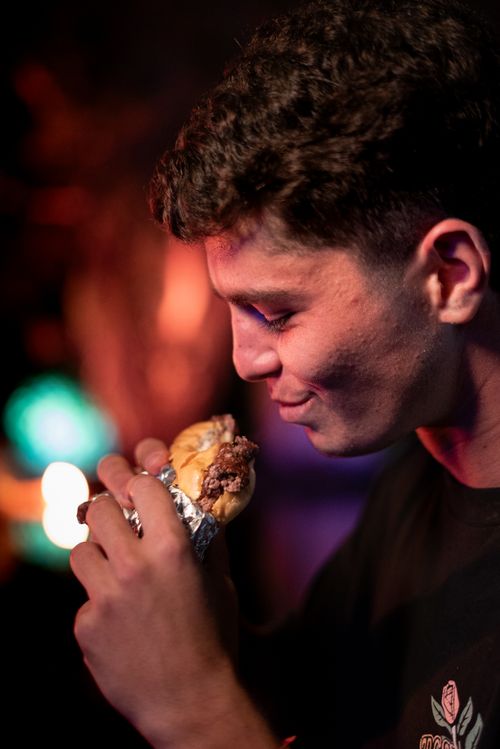 Free A young man savoring a delicious burger wrapped in foil at a vibrant night setting. Stock Photo