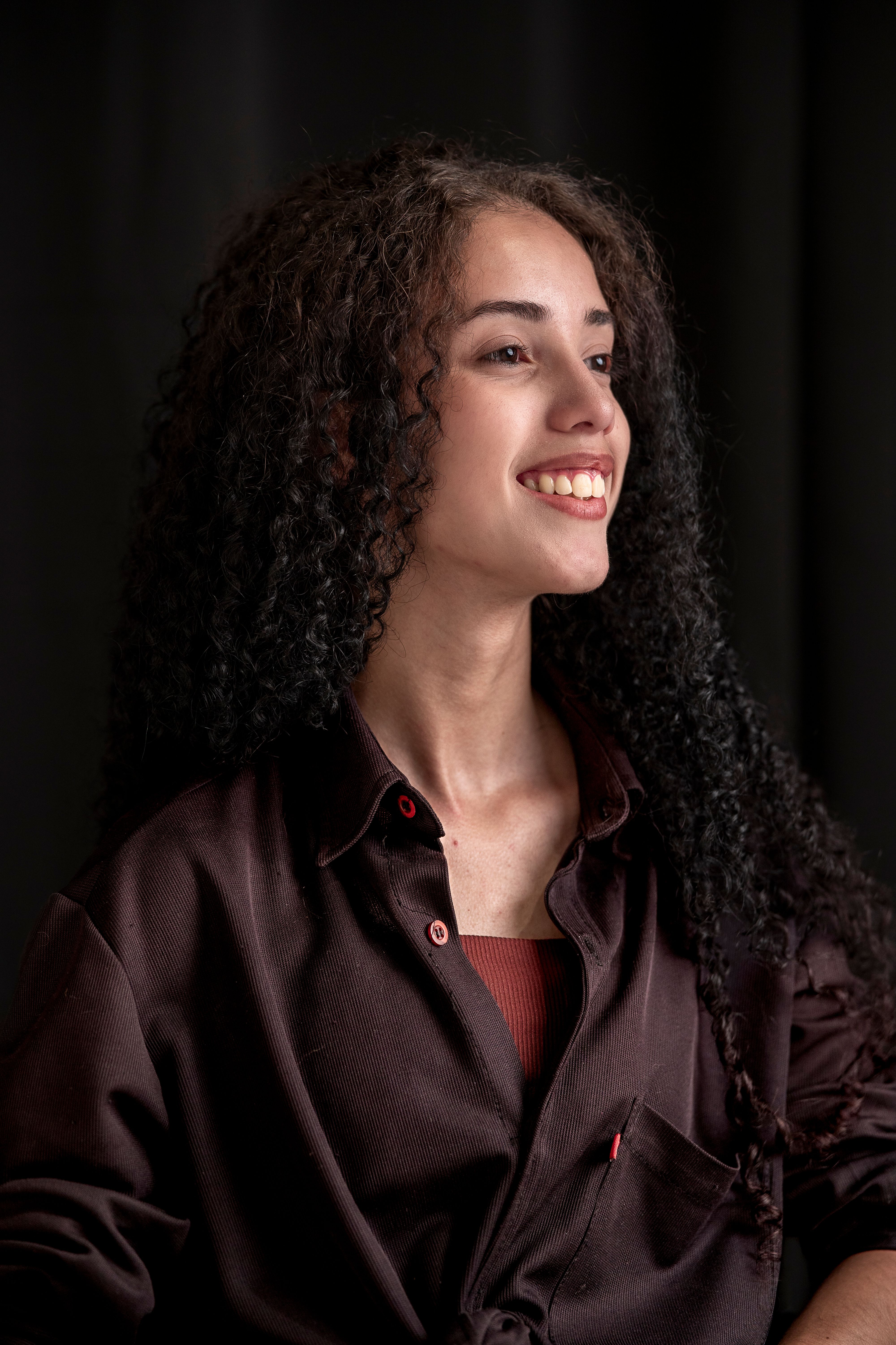 A candid portrait of a smiling woman with curly dark hair against a dark background.