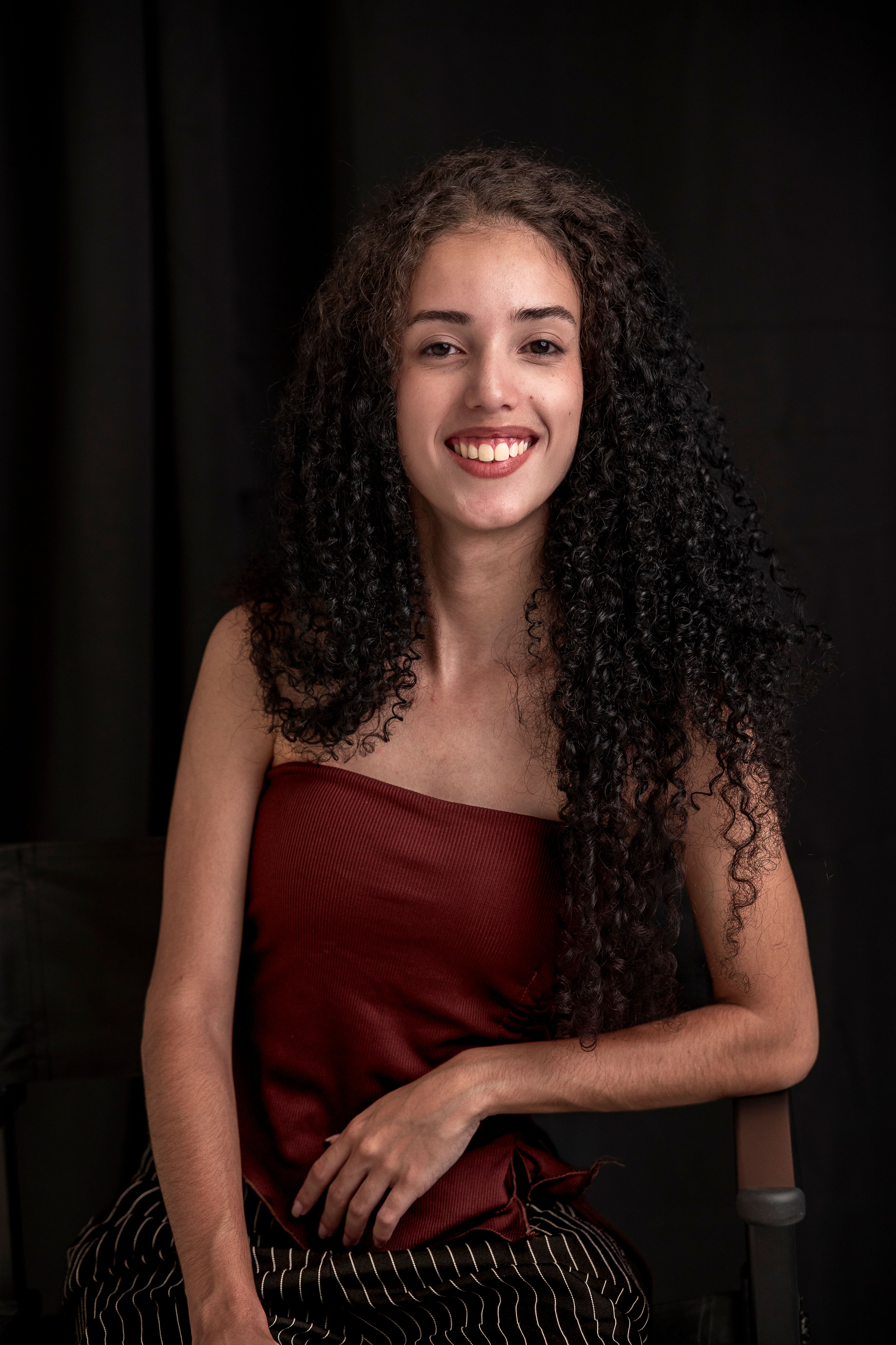 Portrait of a young woman smiling with long curly hair, seated against a dark background.