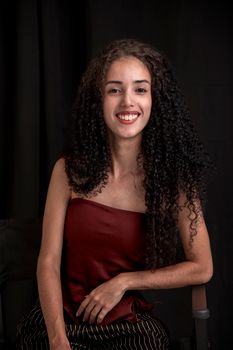 Portrait of a young woman smiling with long curly hair, seated against a dark background.