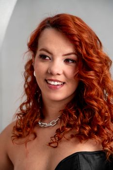 Portrait of a smiling woman with curly red hair, wearing elegant jewelry in a studio setting.