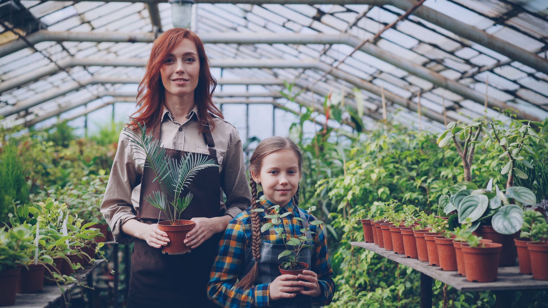 Mother and daughter smiling in a greenhouse with potted plants.