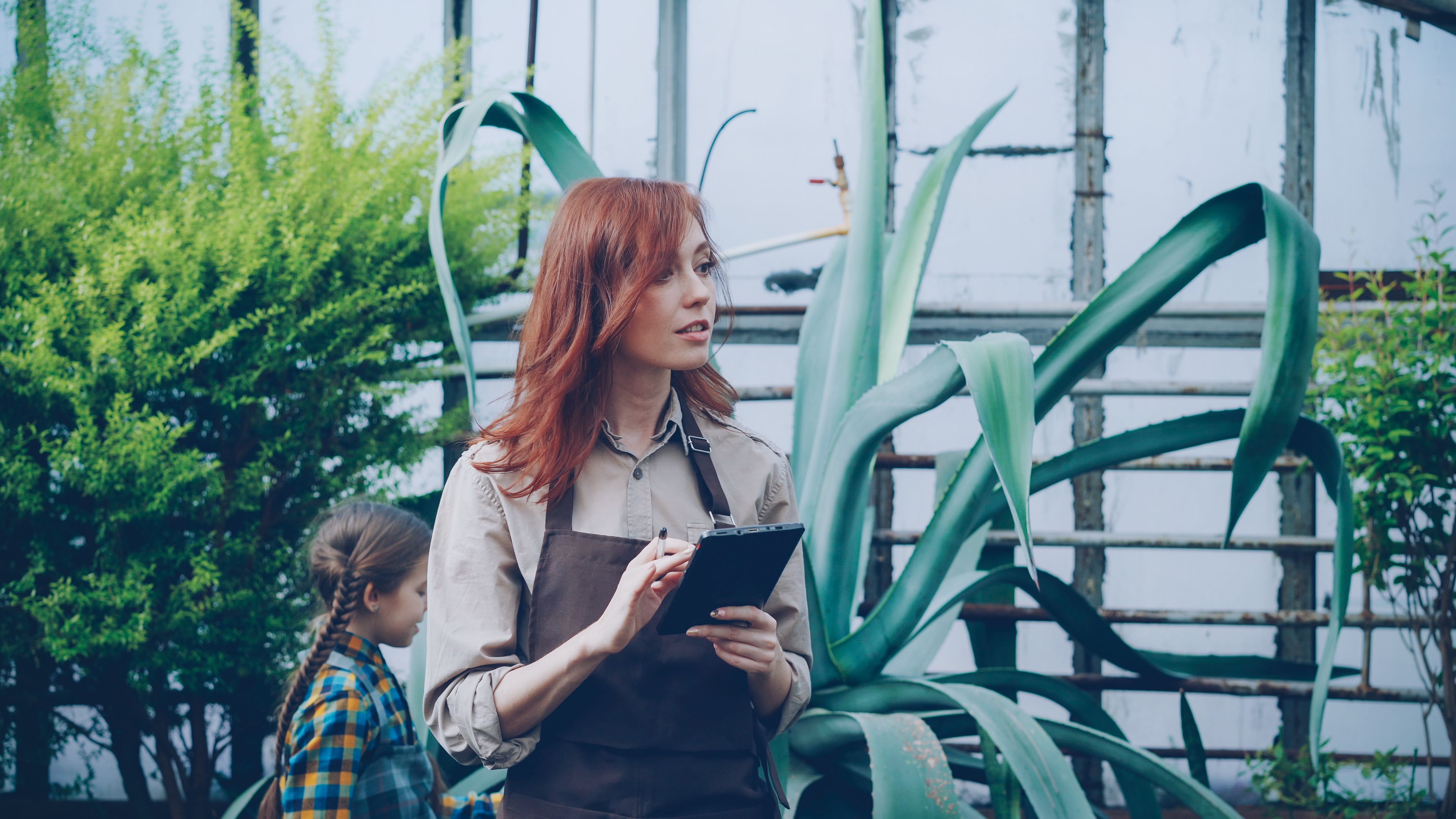 Female gardener with tablet in greenhouse, surrounded by lush agave plants and greenery.