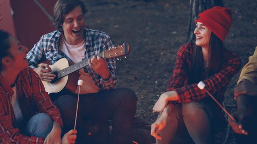 Free Group of friends enjoying a night campfire, playing guitar, and roasting marshmallows outdoors. Stock Photo