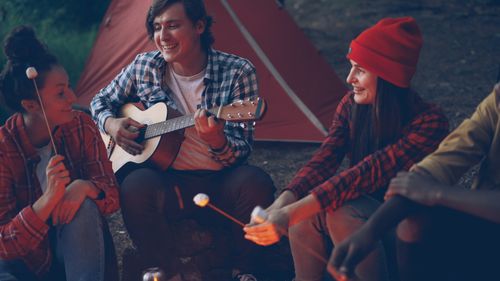 Free Group of friends having fun at a campfire, playing guitar and roasting marshmallows in the evening. Stock Photo