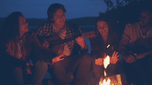 Free Group of friends relaxing by a campfire with music under a night sky. Stock Photo