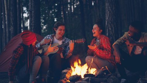 Free Four friends enjoy playing guitar and singing around a campfire in the woods. Stock Photo