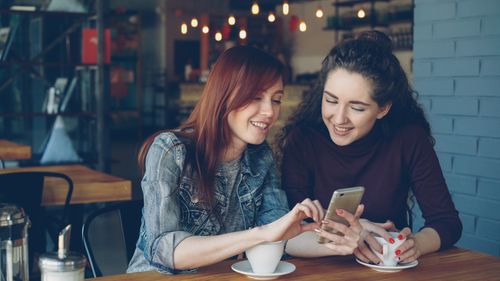 Free Two women share a moment over coffee in a trendy cafe, looking at a smartphone. Stock Photo