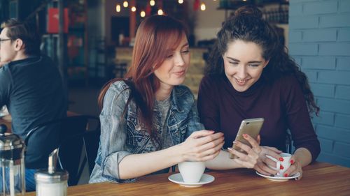Free Two young women sipping coffee and looking at a smartphone inside a cozy café. Stock Photo