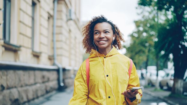 Smiling woman in yellow raincoat walking outdoors while listening to music.