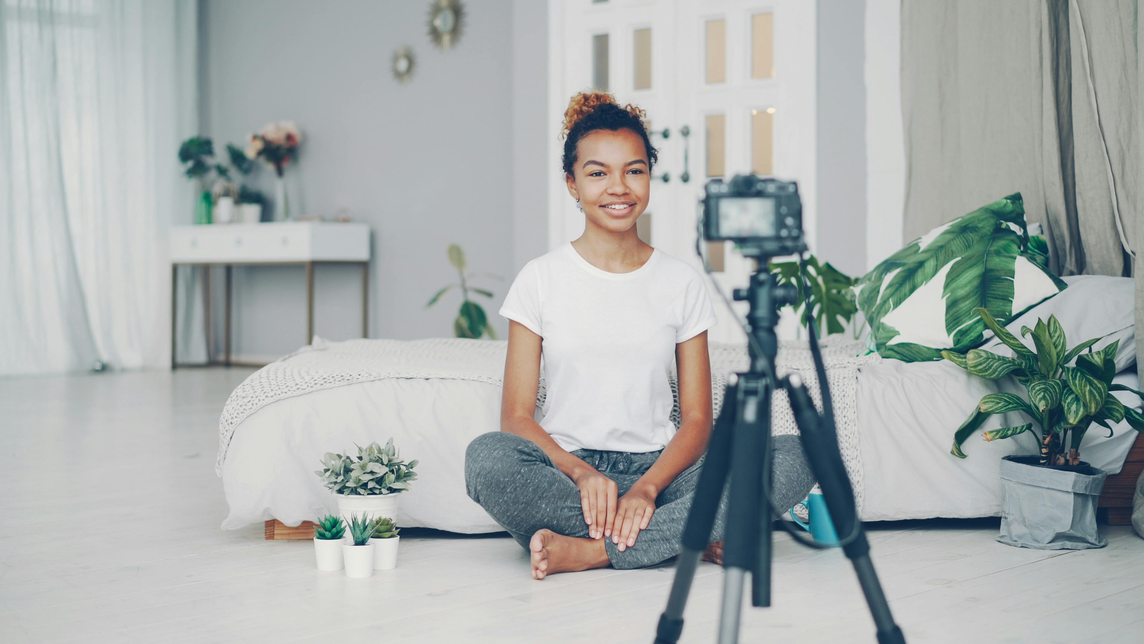 Smiling woman filming a vlog in cozy indoor setting with plants and soft lighting.