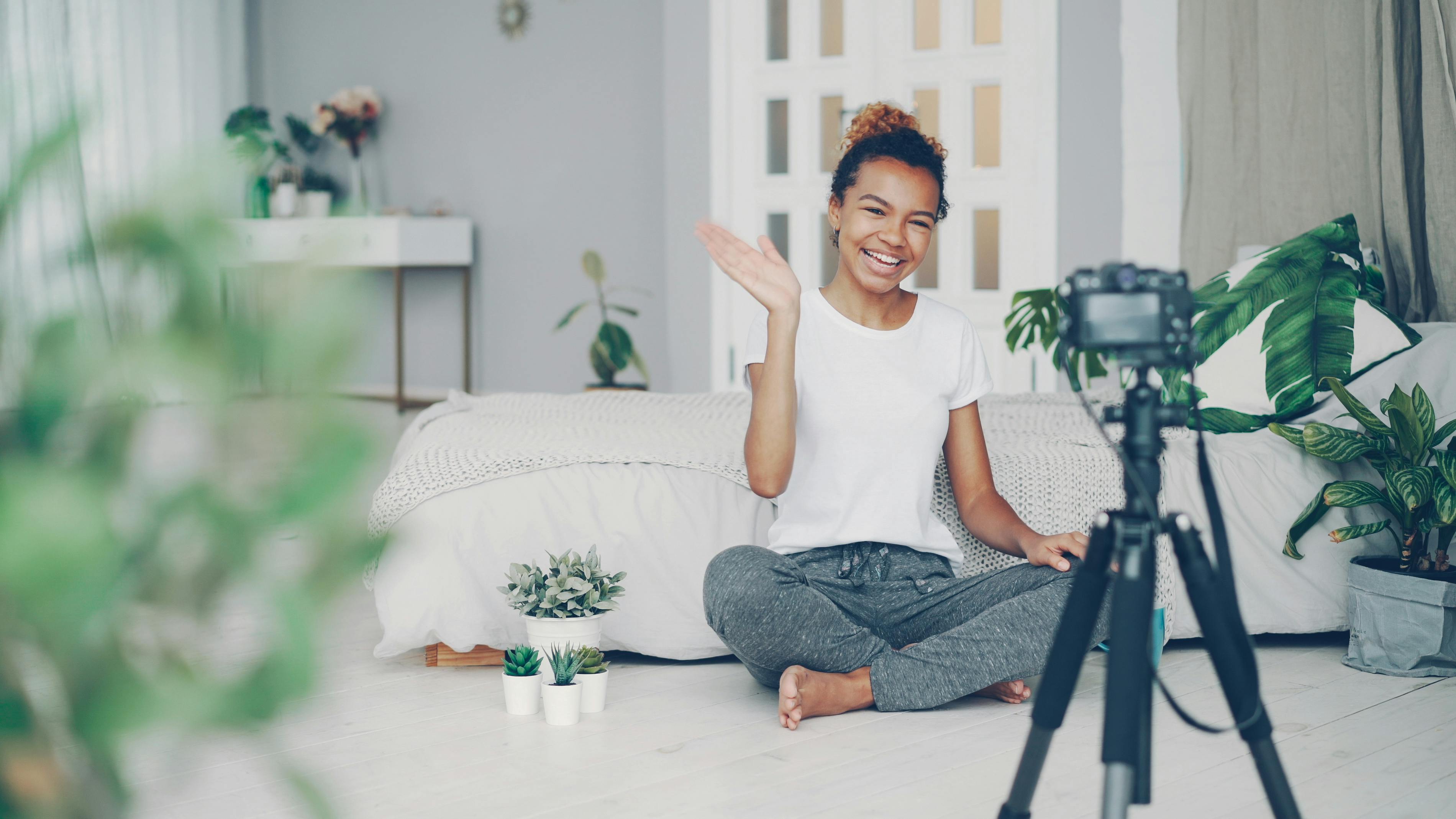 Smiling woman waves at camera while recording a vlog in a stylish bedroom setting.