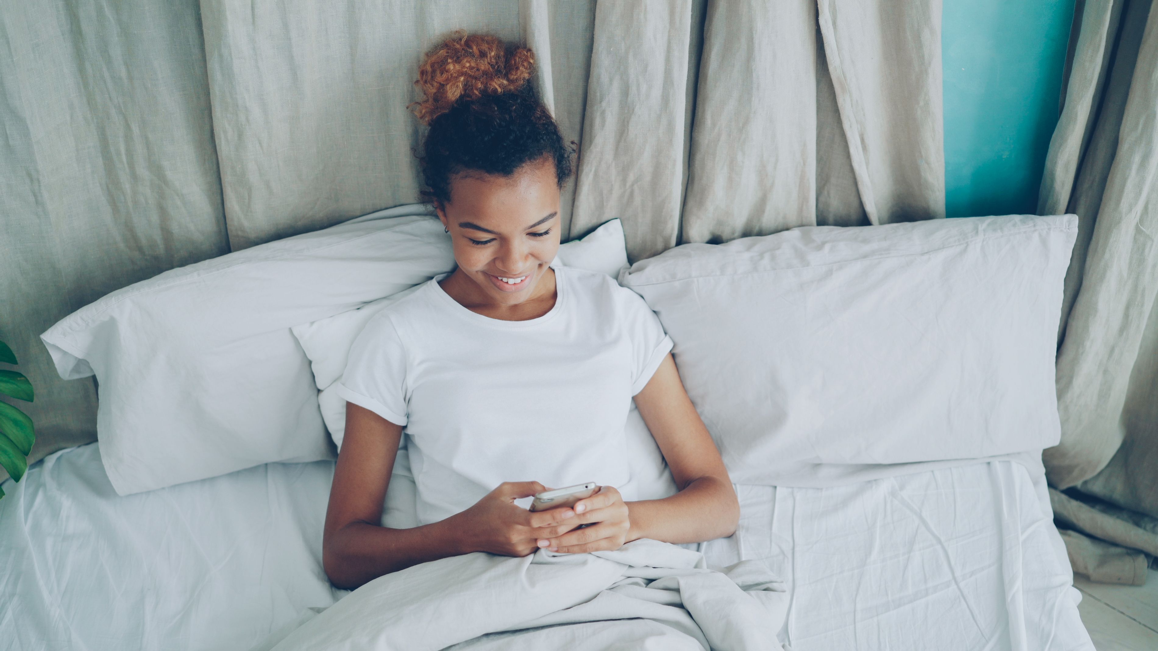 Young woman smiling while using her smartphone in a comfortable bedroom setting.