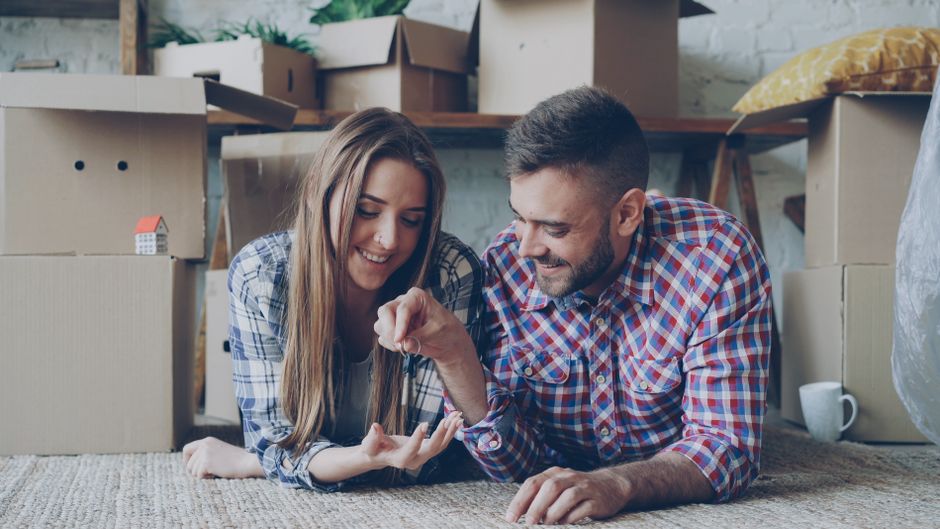 Joyful couple laying on floor surrounded by moving boxes, holding keys to their new home.