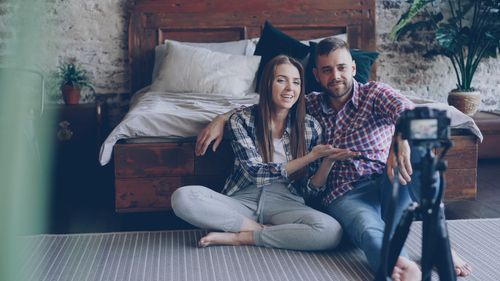 Free A young couple sits together in a cozy bedroom, filming a vlog with a camera on a tripod. Stock Photo