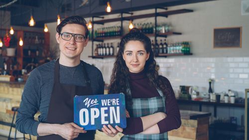 Free Friendly café owners holding an 'Open' sign welcoming customers into their modern coffee shop. Stock Photo