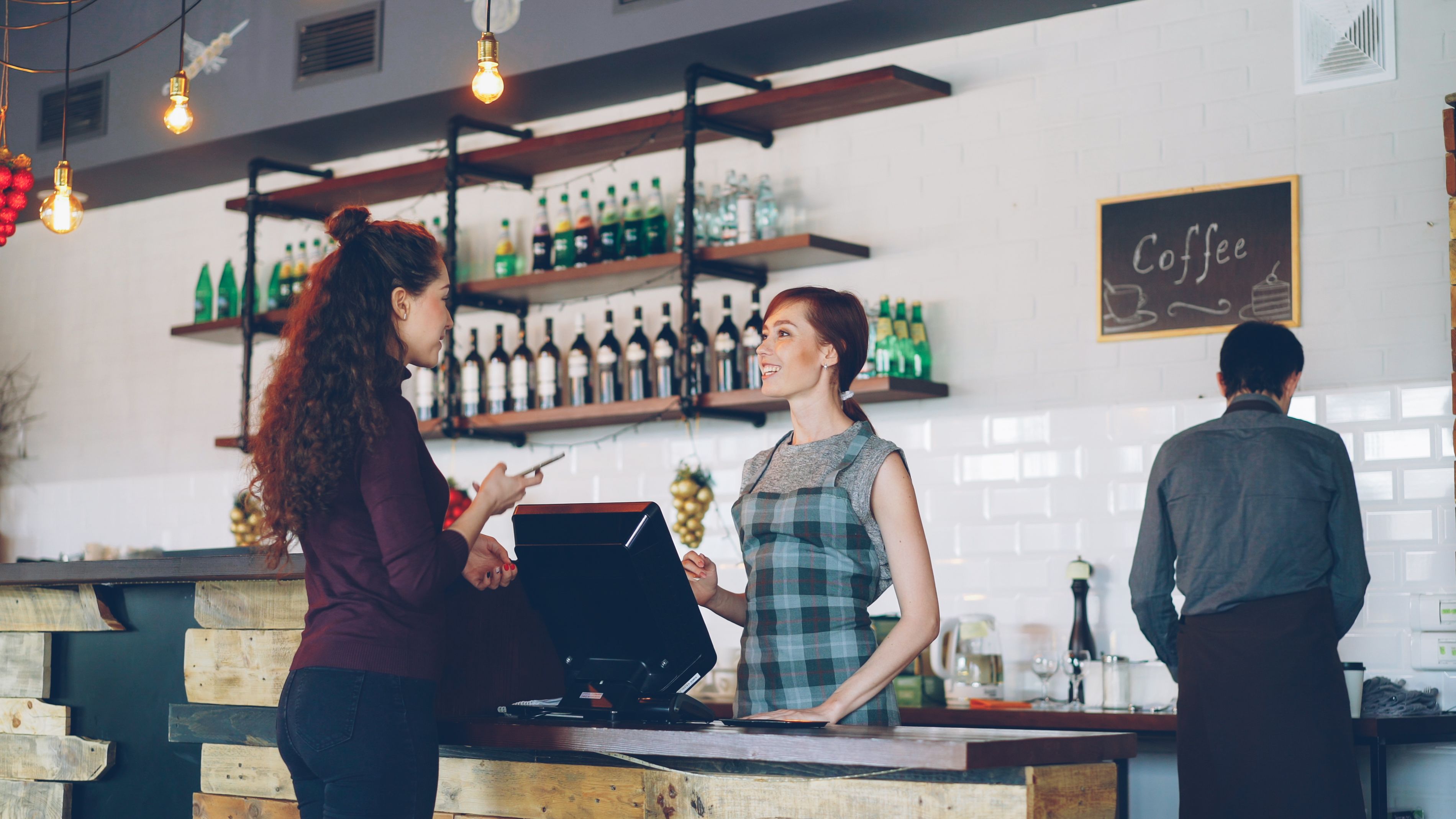 Two women engaged at a coffee shop counter with a barista working in the background.
