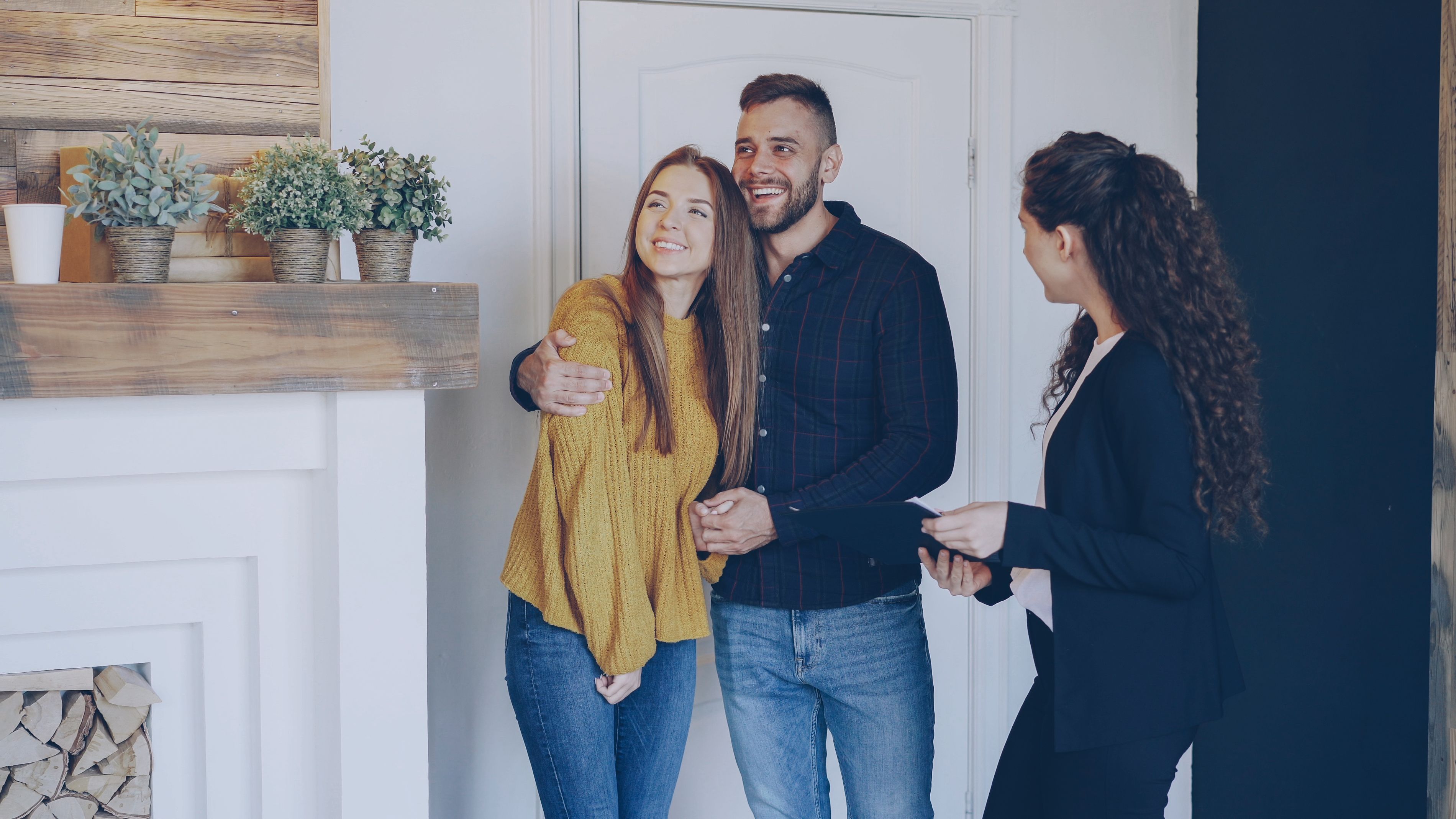 A happy couple embraces in a bright, modern home, interacting with a woman. Features a fireplace with plants and a white door.