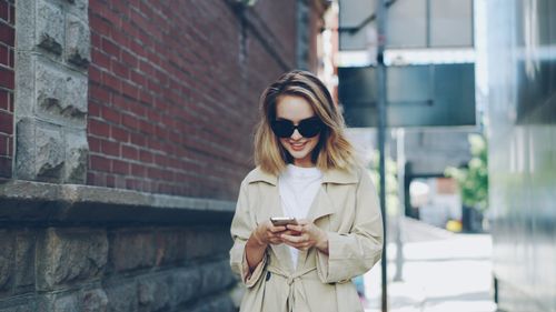Free Stylish woman with sunglasses and smartphone smiling in an urban setting. Stock Photo