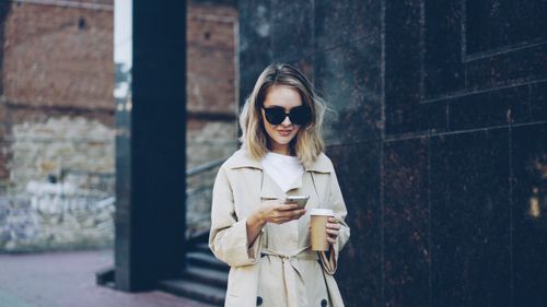Free Young woman in trench coat using smartphone and holding a coffee cup outdoors. Stock Photo