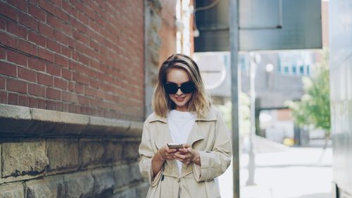 Free Young woman in trench coat texting on smartphone in a sunny urban alley. Stock Photo