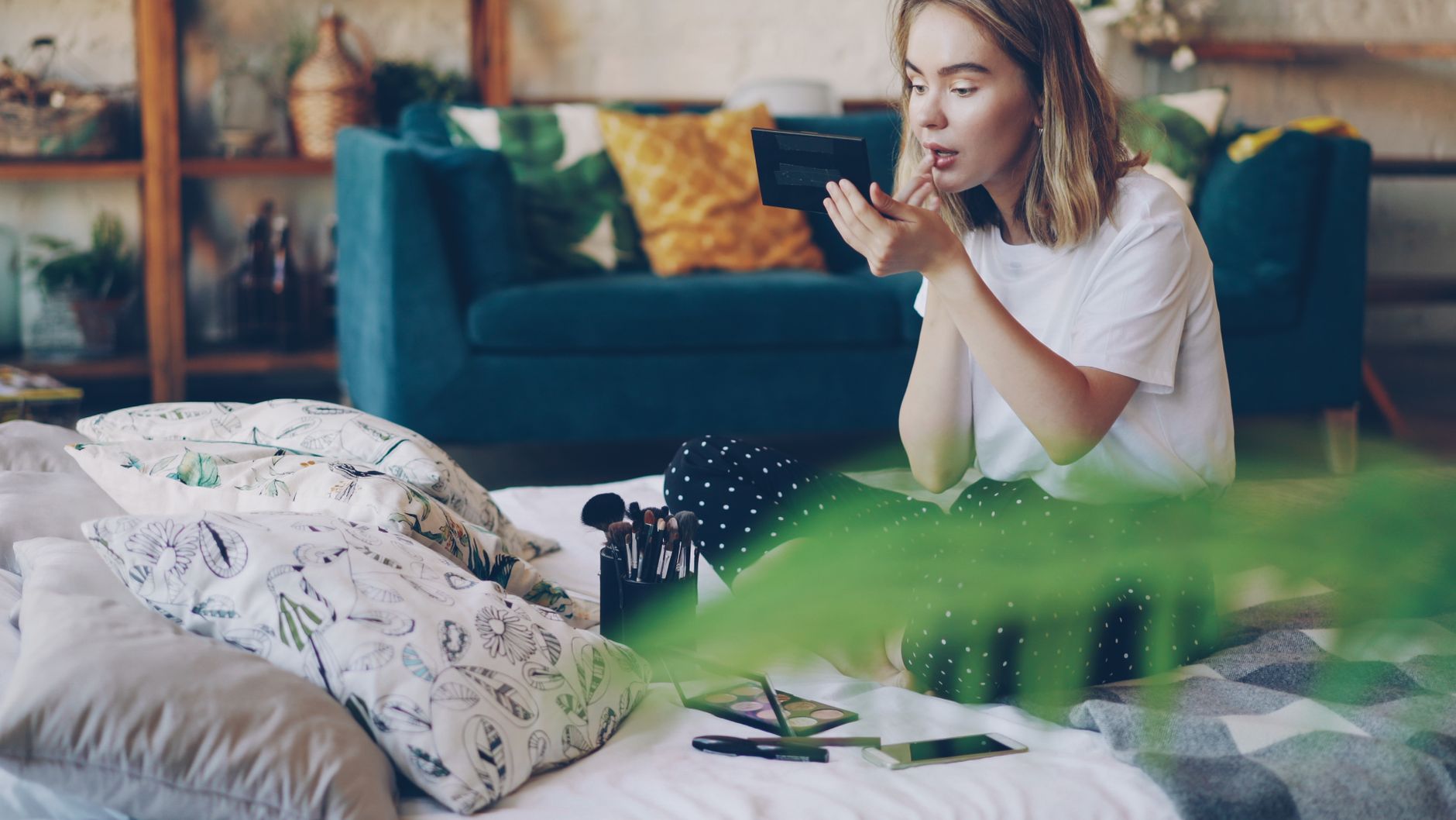 Young adult woman applies makeup sitting on bed in a cozy, stylish room. Ideal for lifestyle themes.
