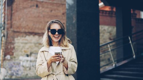 Free Smiling woman in sunglasses listening to music on smartphone outdoors. Stock Photo