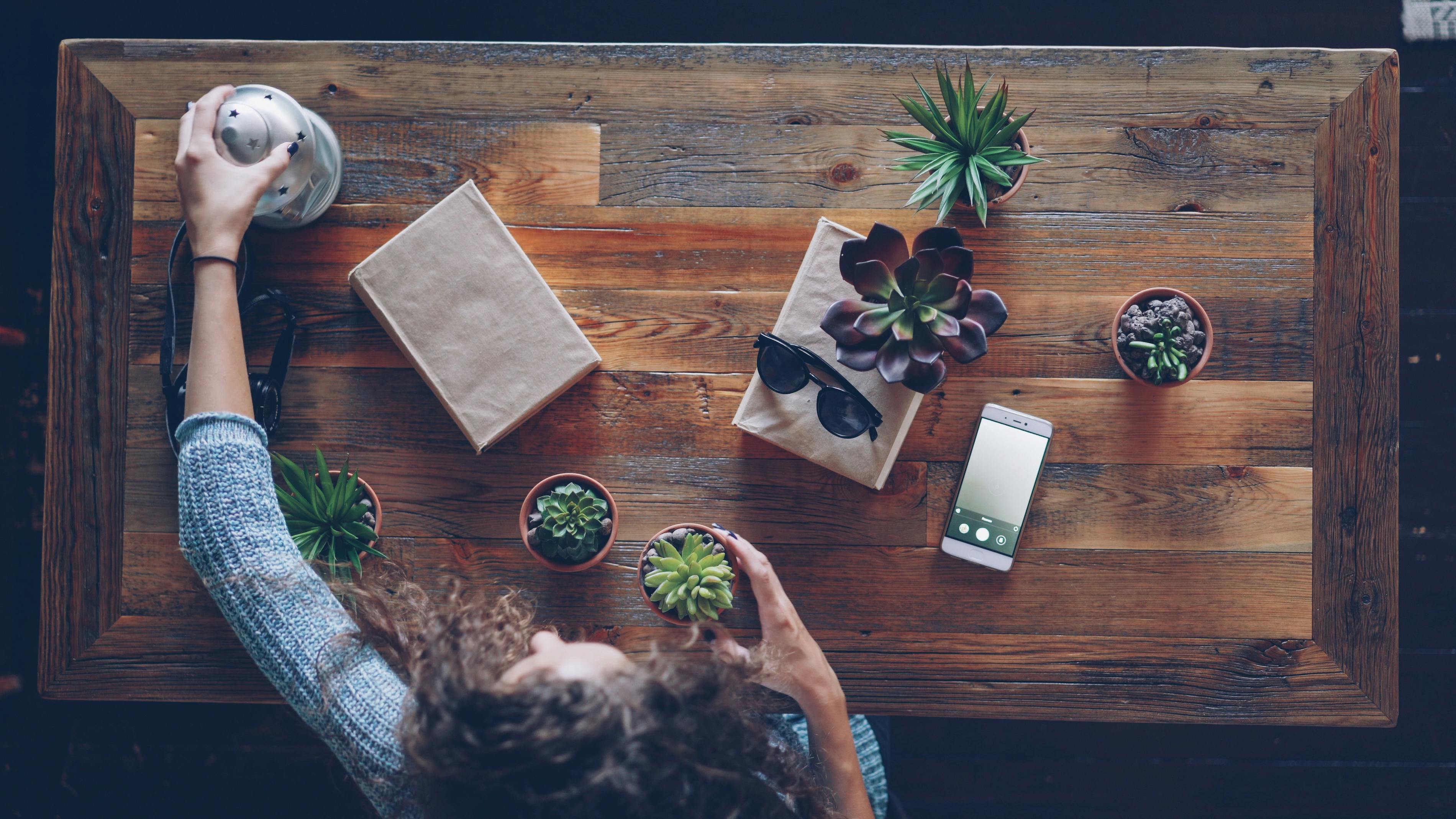 Overhead view of a cozy wooden table setup featuring plants, phone, and accessories.