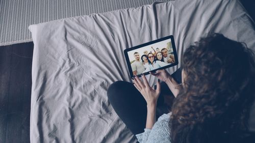 Free Woman sitting on bed using tablet to view a group selfie indoors. Stock Photo