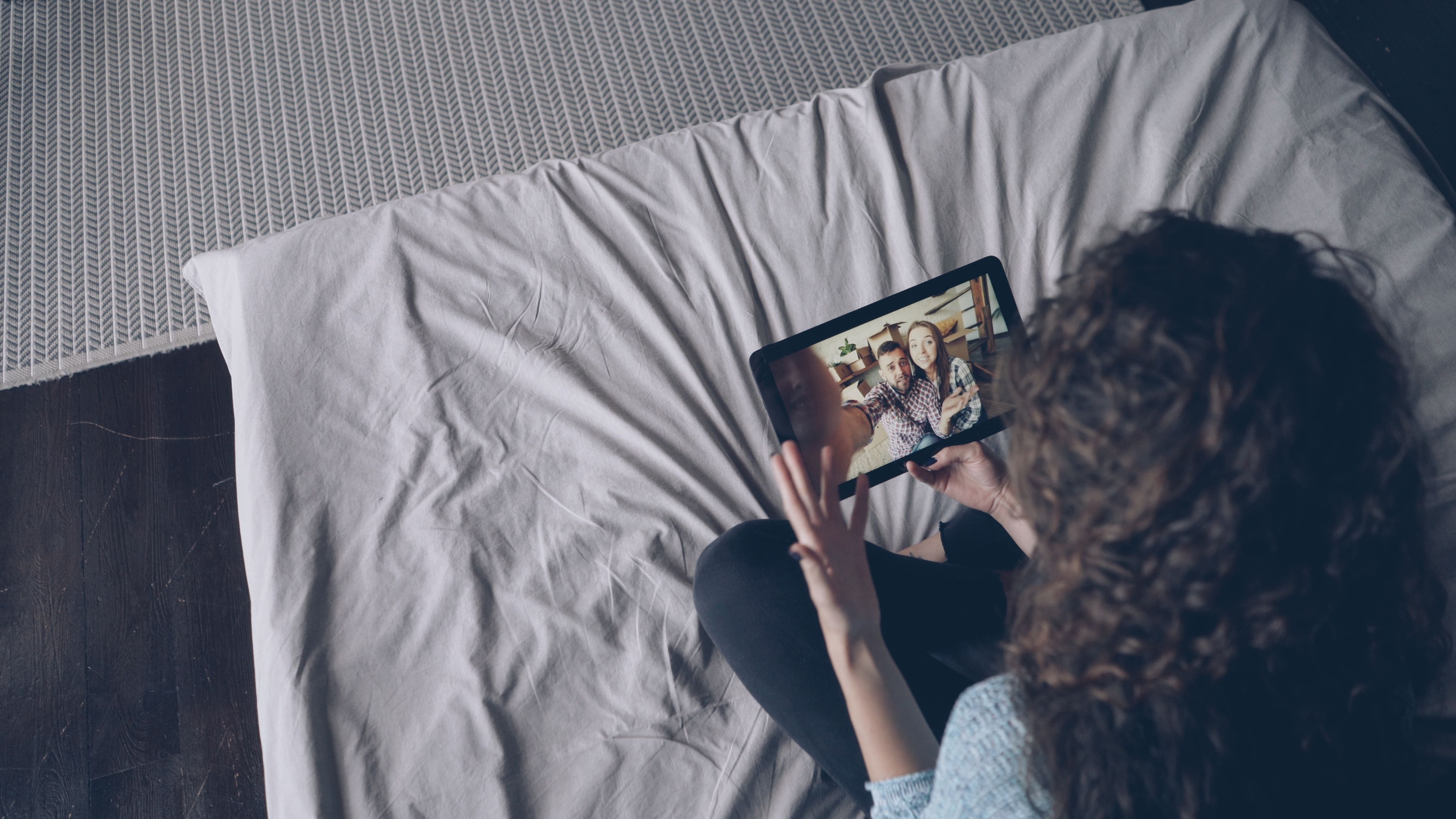 A woman uses a tablet for a video call with friends while sitting on a bed indoors.