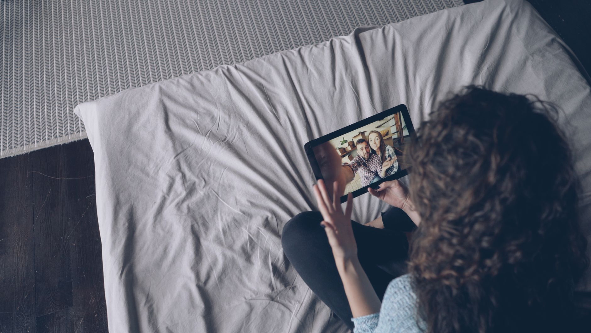 A woman uses a tablet for a video call with friends while sitting on a bed indoors.