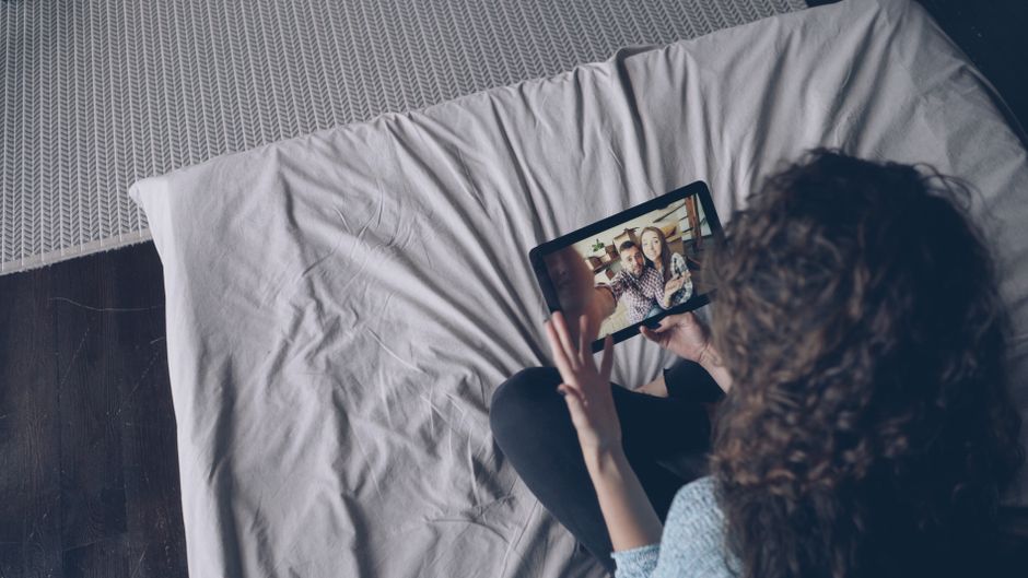 A woman uses a tablet for a video call with friends while sitting on a bed indoors.