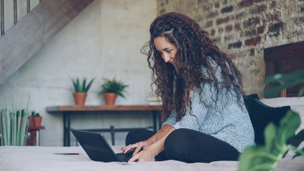 A young woman sits comfortably on her bed using a laptop in a stylish, cozy room.