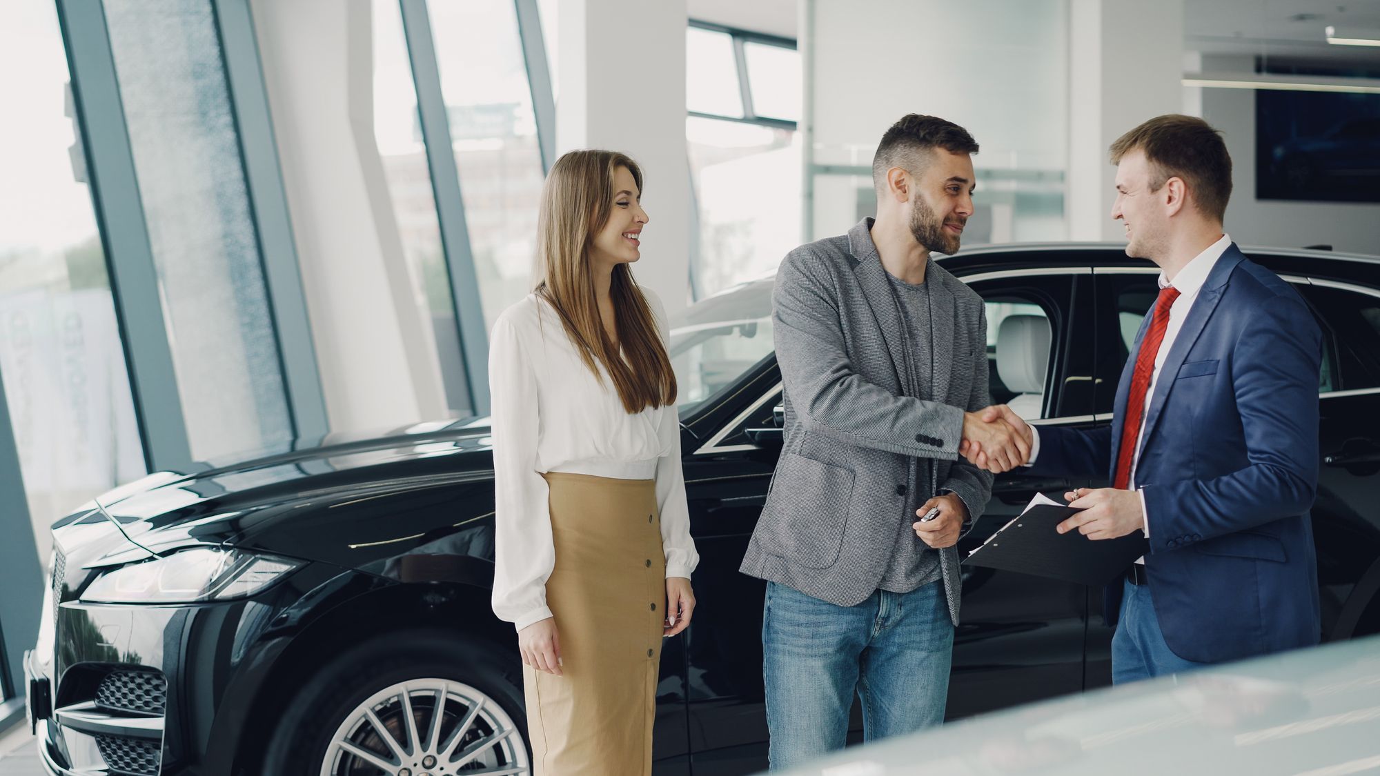 A car dealer shaking hands with a couple in front of a vehicle in a showroom