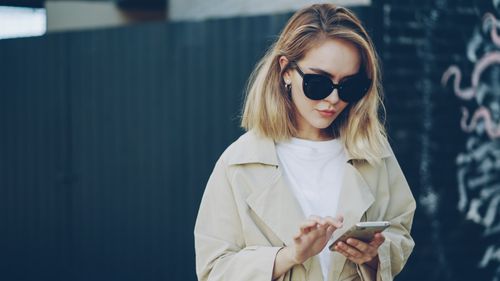 Free Trendy woman in sunglasses and trench coat using smartphone outside. Stock Photo