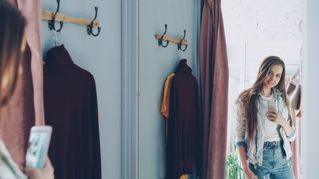Young woman taking a selfie in a dressing room with outfits hanging on hooks.