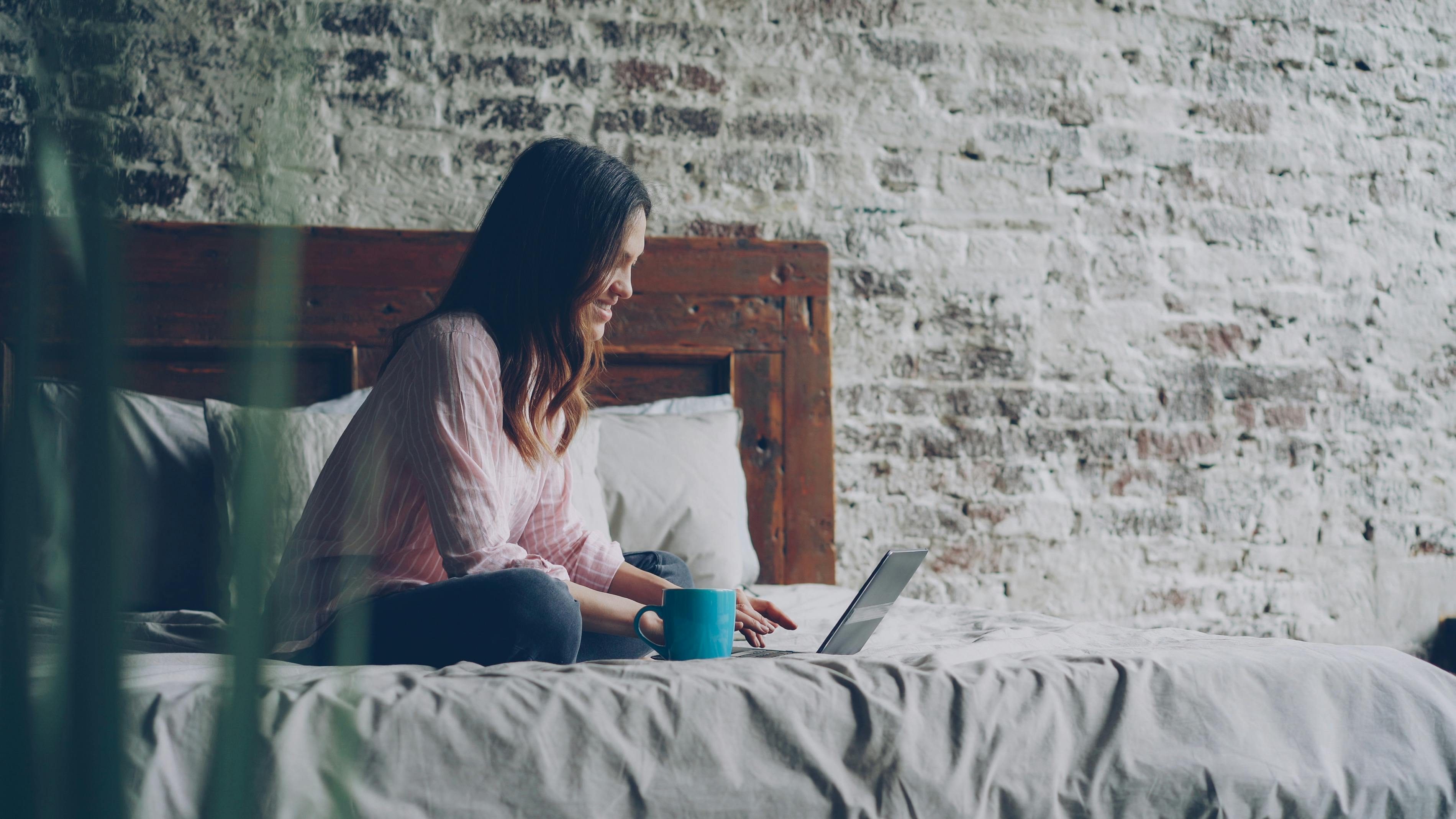 A woman sits comfortably on a bed using a laptop, enjoying a relaxed work-from-home setup.