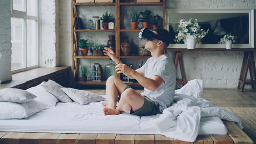 Free Young man enjoying virtual reality headset on bed in cozy room. Stock Photo